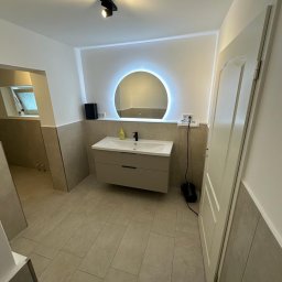 AB Construction - Jasna łazienka z podwieszaną szafką umywalkową, okrągłym lustrem LED z efektem halo, beżowymi płytkami na podłodze i do połowy ścian, oraz czarnym oświetleniem szynowym.