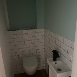 AB Construction - Nowoczesna toaleta z białymi płytkami typu subway na ścianach, zieloną farbą, kompaktową umywalką z czarną baterią i drewnianą podłogą.
