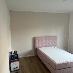 AB Construction - Jasnobeżowy pokój z drewnianą podłogą, różowym tapicerowanym łóżkiem z zagłówkiem oraz pudełkiem odkurzacza Tineco w rogu.