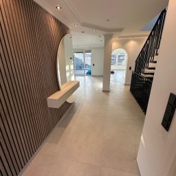 AB Construction - Nowoczesny hol z drewnianymi panelami ściennymi, okrągłym lustrem i minimalistyczną konsolą. Jasne płytki podłogowe, białe ściany, ozdobne listwy sufitowe i czarna balustrada schodów.