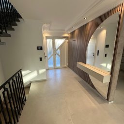 AB Construction - Elegancki przedpokój z beżowymi płytkami, schodami z czarną, geometryczną balustradą oraz ścianą z drewnianych lameli z owalnym lustrem i półką.