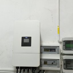 SzychTech Energy - Biały inwerter fotowoltaiczny Deye z wyświetlaczem, zamontowany na teksturowanej ścianie, obok rozdzielnic PV DC i AC z widocznym okablowaniem.