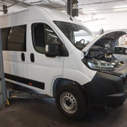 Warsztat samochodowy EkoWodrol - Biały furgon z otwartą maską, częściowo uniesiony na szarym podnośniku hydraulicznym w warsztacie samochodowym, z widocznymi elementami silnika.