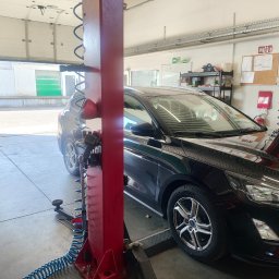 Warsztat samochodowy EkoWodrol - Ciemny samochód osobowy na czerwonym podnośniku hydraulicznym w warsztacie, z otwartą bramą garażową i widocznym wężem pneumatycznym.