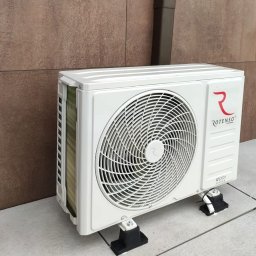 Paweł Stawicki Electric Energy - Biała jednostka zewnętrzna klimatyzacji Rotenso Roni Series, zamontowana na czarnych podstawkach antywibracyjnych na płytkach, przy brązowej ścianie z rynną.