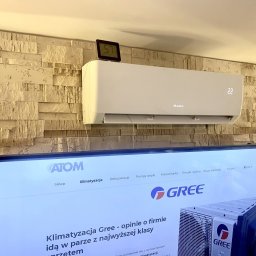 Paweł Stawicki Electric Energy - Biała jednostka klimatyzacji GREE z cyfrowym wyświetlaczem '22', zamontowana na beżowej ścianie z kamienia dekoracyjnego. Nad nią zegar '57', a poniżej ekran TV z ofertą GREE.