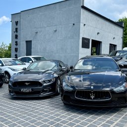 FHU VERSUS SP. Z O.O. - Lśniące czarne Maserati Ghibli i ciemnoszary Ford Mustang zaparkowane przed nowoczesnym budynkiem z pionowym napisem VERSUS, na tle kostki brukowej.
