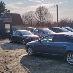 Skup Aut Elbląg El-Fura - Używane samochody, w tym granatowe Audi A6 i Volvo S40, zaparkowane na nierównym placu obok kontenerowego biura z napisem 'AUTO HANDEL' w słoneczny dzień.