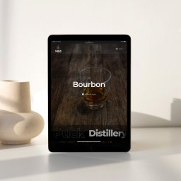 EnterSpace - Tablet wyświetlający stronę internetową Puck Distillery z dużą grafiką szklanki bourbonu na ciemnym drewnie, obok beżowy wazon.