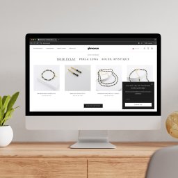 EnterSpace - Monitor wyświetlający sklep internetowy glamourya.pl z biżuterią (bransoletki, kolczyki, naszyjniki) z kolekcji Noir Éclat i pop-up newslettera, na drewnianym biurku.