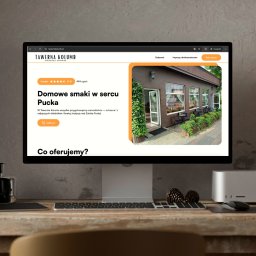 EnterSpace - Strona internetowa Tawerny Kolumb na monitorze, prezentująca ofertę 'Domowe smaki w sercu Pucka' ze zdjęciem restauracji. Na drewnianym biurku klawiatura, mysz i szyszki.