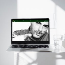 EnterSpace - Srebrny laptop wyświetlający czarno-białe zdjęcie mężczyzny wskazującego palcem, z napisem 'August Kaiser' na stronie akaiser.pl. Obok szklanka wody i szklana butelka, tło z geometrycznymi cieniami.