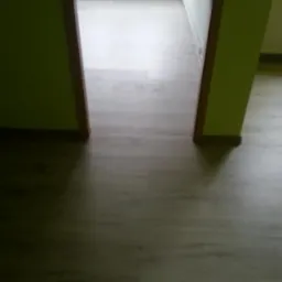 Widok z perspektywy podłogi na świeżo położone panele podłogowe w odcieniach szarości, z widocznym fragmentem zielonej ściany i futryny drzwi.