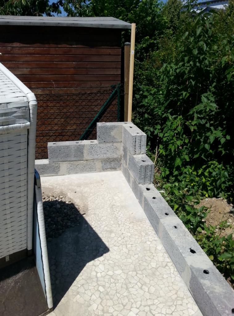 Budowa niskiego murku z szarych bloczków betonowych na zewnątrz, widoczny fragment białego mebla ogrodowego, brązowy budynek gospodarczy w tle i zieleń.