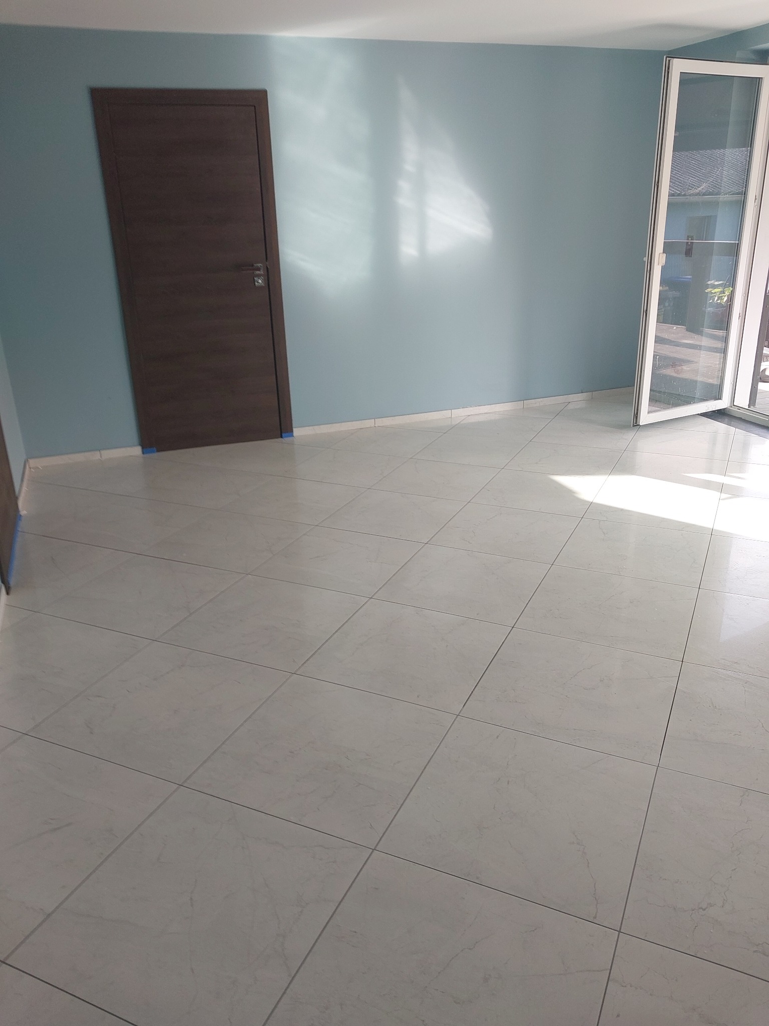 Jasne, kwadratowe płytki podłogowe z delikatnym wzorem marmuru w nowym pokoju z błękitnymi ścianami, brązowymi drzwiami i otwartym oknem balkonowym.