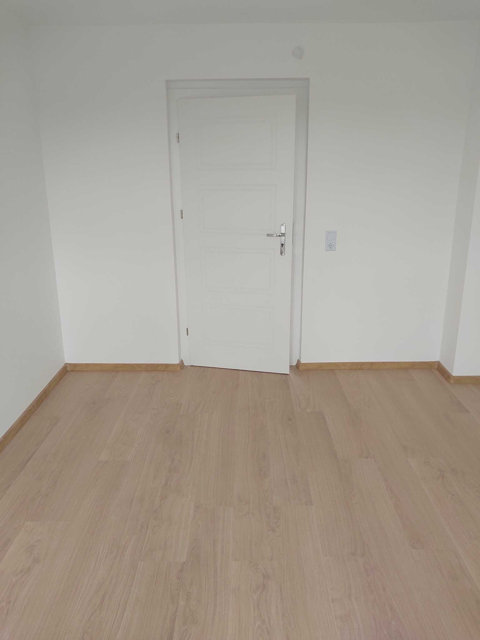 Jasne panele podłogowe w nowym pokoju z białą ścianą i drzwiami. Widoczne listwy przypodłogowe. Minimalistyczne wnętrze po remoncie.