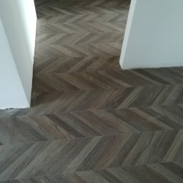 Usługi remontowe - Podłoga z paneli lub płytek imitujących ciemne drewno, ułożona w precyzyjny wzór jodełki, biegnąca przez białe ościeżnice do kolejnego pomieszczenia.