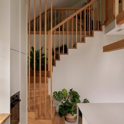 DO.NUS STUDIO Dominika Augustynek - Nowoczesne wnętrze z jasnymi, drewnianymi schodami o pionowych balustradach, prowadzącymi na antresolę. Obok minimalistyczna kuchnia i rośliny.