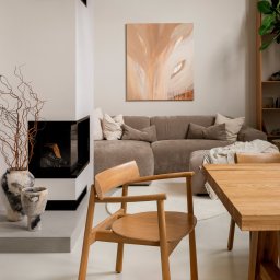 DO.NUS STUDIO Dominika Augustynek - Nowoczesny salon z jadalnią: beżowa sofa modułowa, drewniany stół, krzesło, narożny kominek z czarnymi akcentami i abstrakcyjny obraz w ciepłych barwach.