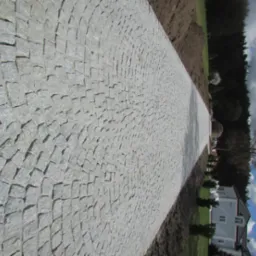 Ścieżka z jasnoszarej kostki, ułożona w łukowaty wzór, prowadząca wzdłuż trawnika i domu w słoneczny dzień.