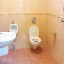 Jasna łazienka z beżowymi płytkami, widoczna umywalka z baterią, pisuar, toaleta z otwartą klapą oraz grzejnik drabinkowy, z tubą silikonu przy umywalce.