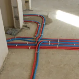 Podłogowe rozprowadzenie instalacji hydraulicznej z rur PEX w kolorze czerwonym i niebieskim, przymocowanych białymi uchwytami do surowej posadzki, widoczne fragmenty ścian w stanie deweloperskim.