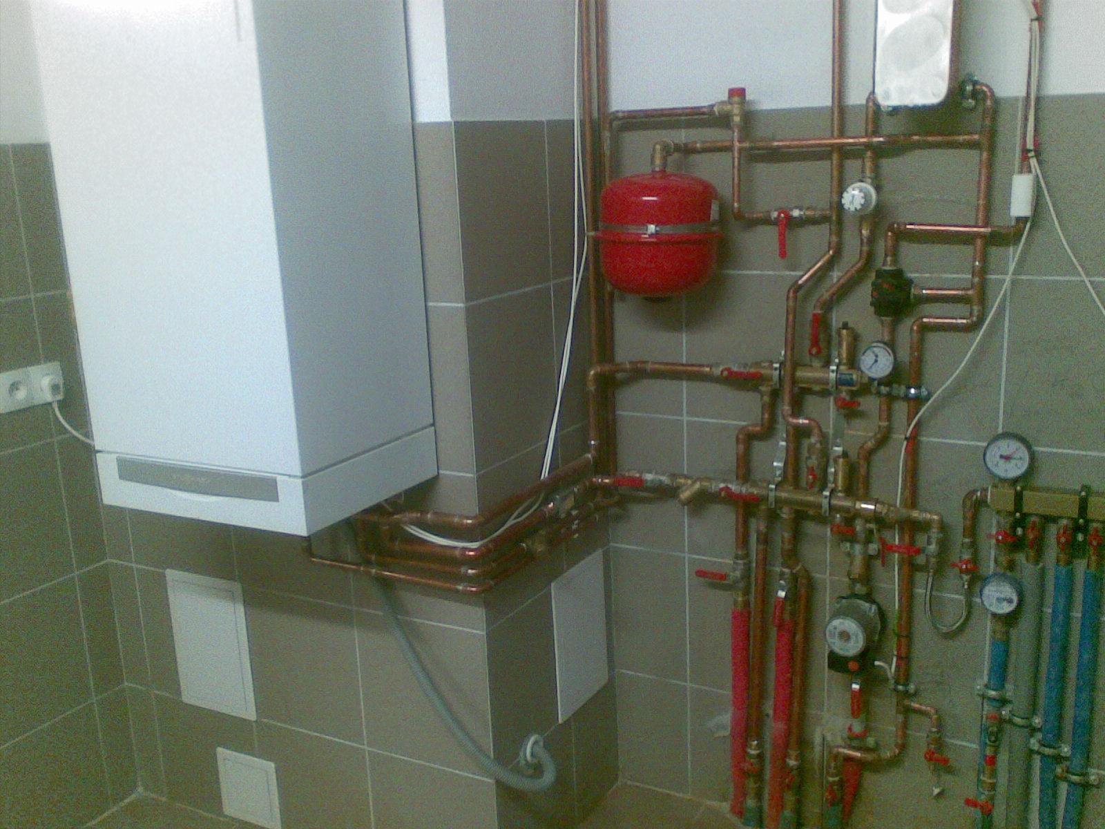 Biały piec grzewczy wiszący na ścianie z szarymi kafelkami, z widoczną instalacją hydrauliczną z miedzianych rur, czerwonym zbiornikiem wyrównawczym i manometrami.