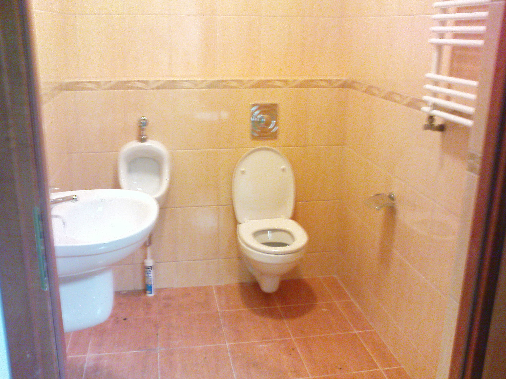 Jasna łazienka z beżowymi płytkami, widoczna umywalka z baterią, pisuar, toaleta z otwartą klapą oraz grzejnik drabinkowy, z tubą silikonu przy umywalce.