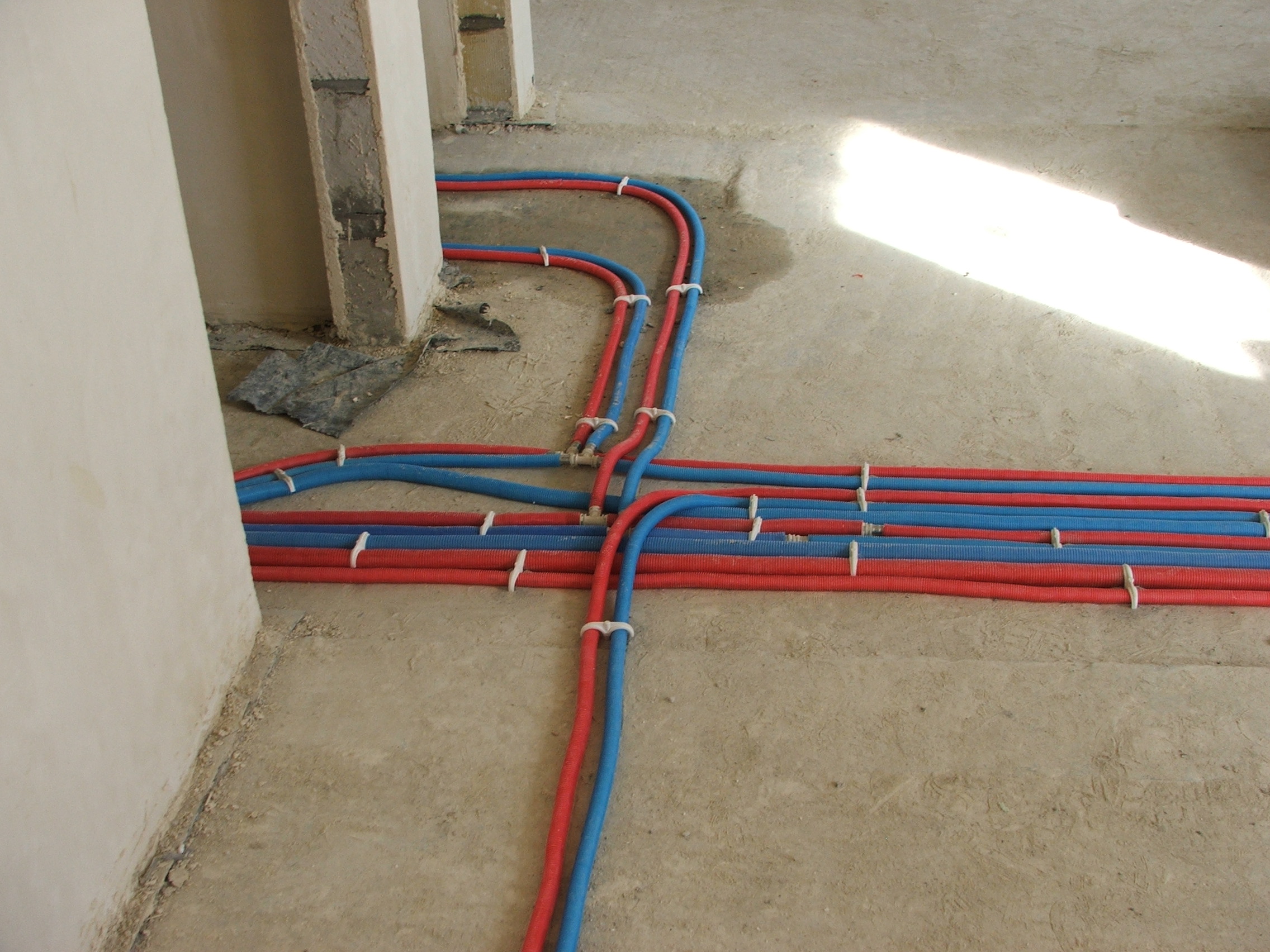 Podłogowe rozprowadzenie instalacji hydraulicznej z rur PEX w kolorze czerwonym i niebieskim, przymocowanych białymi uchwytami do surowej posadzki, widoczne fragmenty ścian w stanie deweloperskim.
