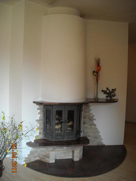 Kotłownia z żeliwnym piecem kaflowym obudowanym kamieniem dekoracyjnym, okrągła obudowa komina, drewniane półki i dekoracje.