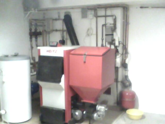 Kotłownia z kotłem na paliwo stałe marki HEITZ, zbiornikiem akumulacyjnym i instalacją hydrauliczną na ścianie, widoczna żółta miska na podłodze.