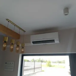 Biała jednostka klimatyzacji zamontowana na ścianie w nowoczesnym wnętrzu, widoczna także designerska lampa wisząca i fragment drewnianej belki stropowej.