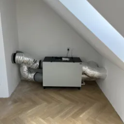 Centrala wentylacyjna z rekuperacją zamontowana na poddaszu, widoczne srebrne, izolowane przewody wentylacyjne, jasne ściany i podłoga w jodełkę.