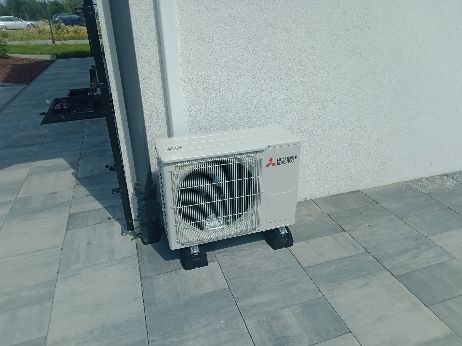 Zewnętrzna jednostka klimatyzacji Mitsubishi Electric zamontowana na czarnych podkładkach antywibracyjnych, ustawiona na szarej kostce brukowej przy białej ścianie budynku, obok rury spustowej.