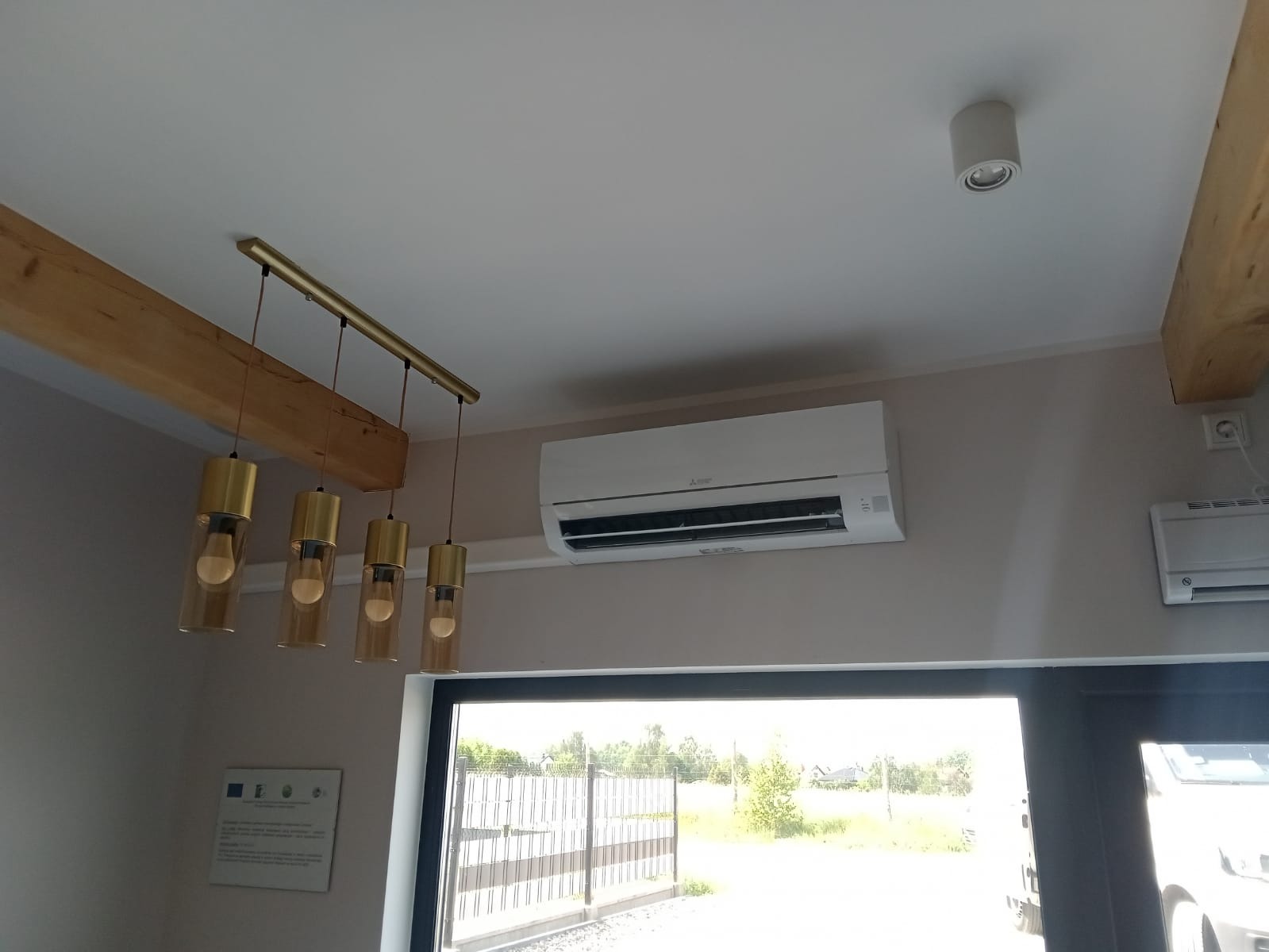 Biała jednostka klimatyzacji zamontowana na ścianie w nowoczesnym wnętrzu, widoczna także designerska lampa wisząca i fragment drewnianej belki stropowej.