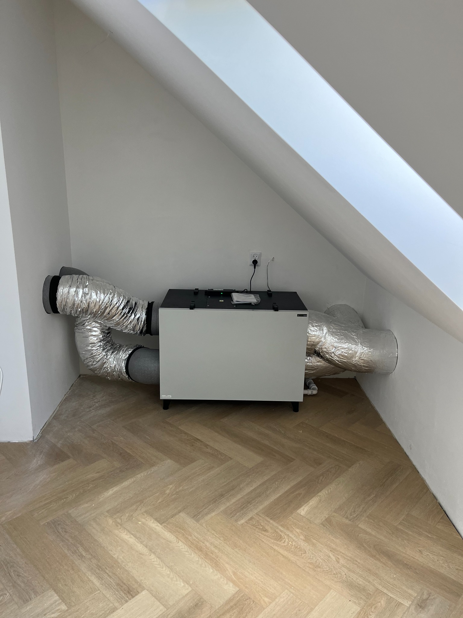 Centrala wentylacyjna z rekuperacją zamontowana na poddaszu, widoczne srebrne, izolowane przewody wentylacyjne, jasne ściany i podłoga w jodełkę.