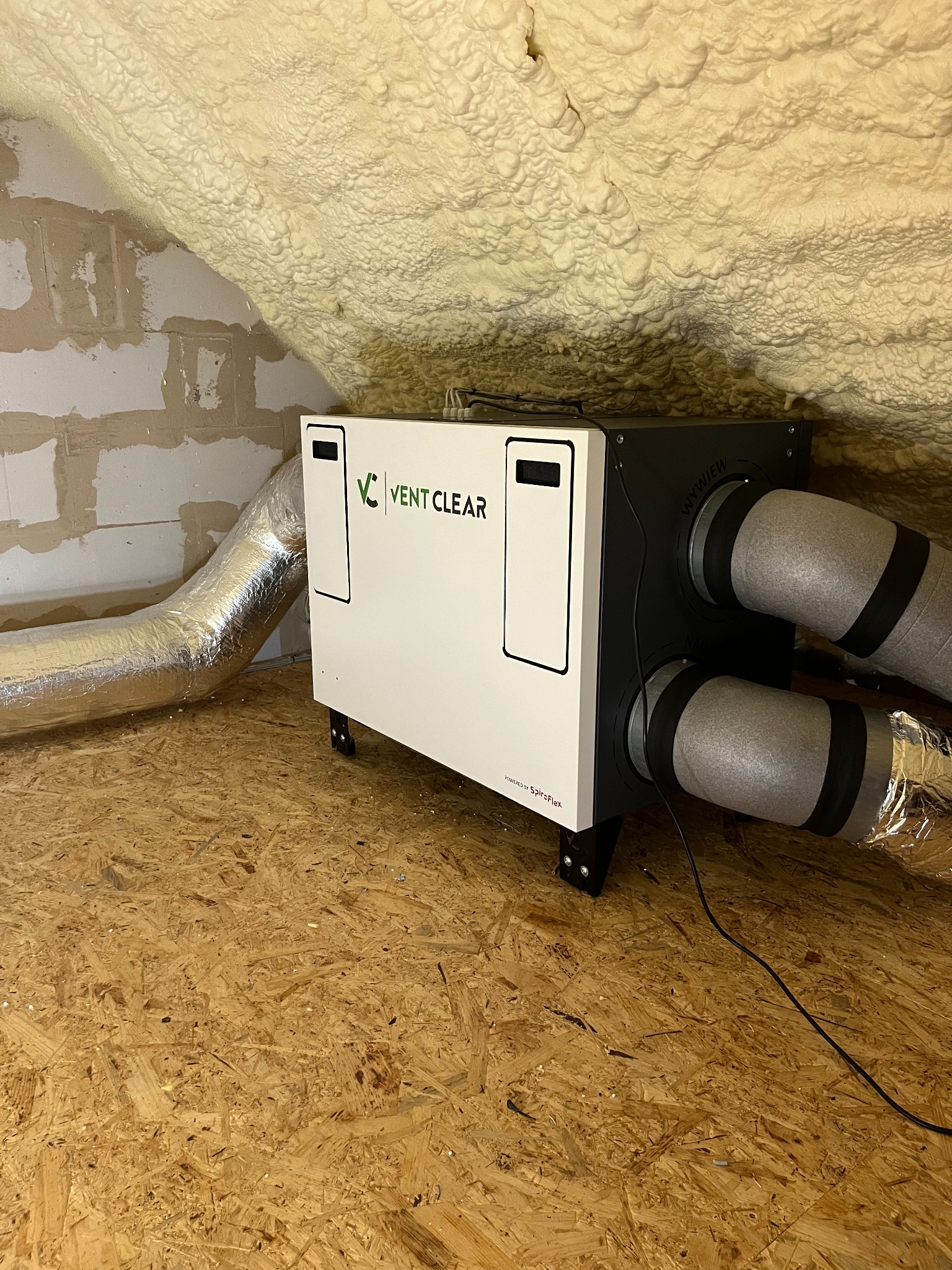 Biała jednostka centralna z logo Vent Clear, podłączona do przewodów wentylacyjnych w zaizolowanym pomieszczeniu na poddaszu z podłogą z płyty OSB.