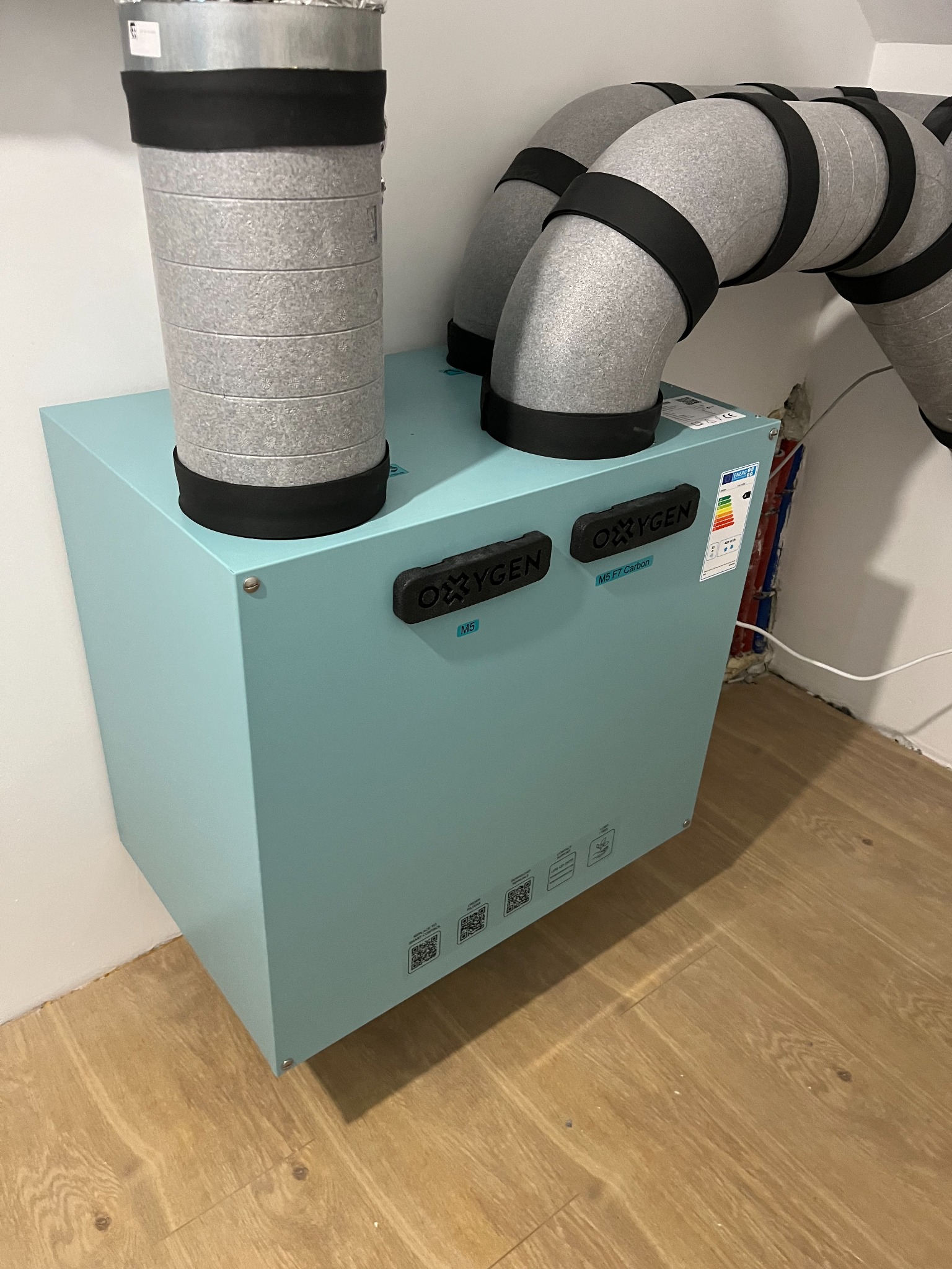 Niebieska jednostka centralna z szarymi przewodami wentylacyjnymi, zamontowana na ścianie w pomieszczeniu z drewnianą podłogą.