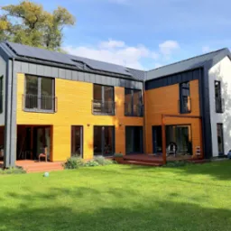 Nowoczesny, dwukondygnacyjny dom z drewnianą elewacją w odcieniach żółci i bieli, z ciemną, metalową dachówką i panelami słonecznymi, otoczony zielonym trawnikiem i drzewami.