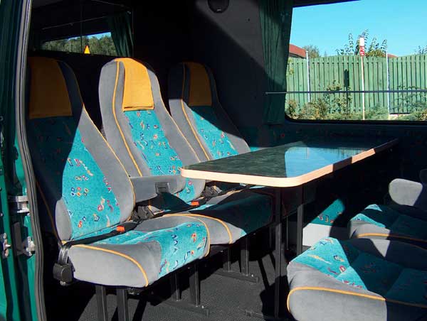 Wnętrze busa pasażerskiego z trzema rzędami siedzeń w kolorze szarym, turkusowym i żółtym, z rozkładanym stolikiem, widok przez okno na zielone ogrodzenie i budynki.