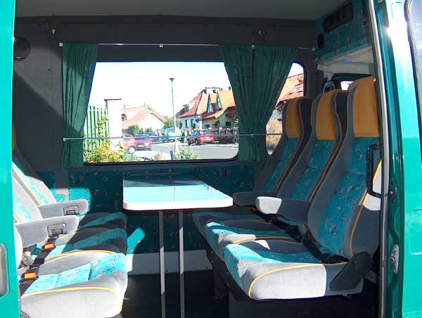 Wnętrze busa z turkusowo-szarymi fotelami, rozkładanym stolikiem i widokiem na ulicę z domami przez okno z zielonymi zasłonami.