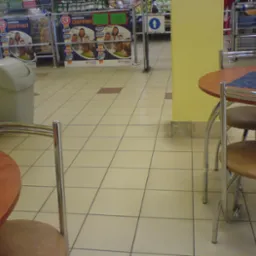 Beżowe płytki podłogowe z ciemnymi fugami w supermarkecie, z fragmentami stolików i krzeseł oraz koszem na śmieci w tle.