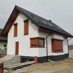 MIG-DACH - Nowy dom z białą elewacją, ciemnym dachem z dachówką, brązową podbitką i rynnami. Widoczne okna dachowe, bariery śniegowe oraz teren budowy z beczką.