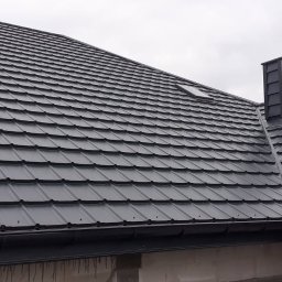 MIG-DACH - Nowoczesny dach z ciemnoszarej blachodachówki z dwoma oknami dachowymi i dopasowanym kominem, widoczny system rynnowy. Poniżej dachu ściana z pustaków.