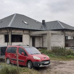 MIG-DACH - Nowo ułożony ciemnoszary dach z dachówek na domu w stanie surowym, z trzema oknami dachowymi i czarnymi rynnami. Przed budynkiem czerwony samochód z przyczepą.