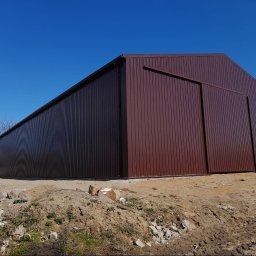 MIG-DACH - Nowoczesny budynek gospodarczy z ciemnobrązowej blachy trapezowej, z dwuspadowym dachem i dużymi przesuwnymi drzwiami, na tle bezchmurnego nieba. Wokół plac budowy.
