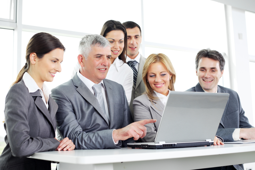 Zespół sześciu osób w garniturach, pochylony nad laptopem na białym stole, omawia strategię marketingową.