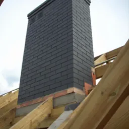 Wysoki komin obłożony grafitową klinkierową cegłą, widoczny na tle konstrukcji dachu w trakcie budowy, z ceglaną bazą i betonowym wykończeniem.