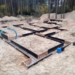 Przygotowanie zbrojenia i szalunków pod fundamenty domu jednorodzinnego na piaszczystym terenie, widoczne wykopane rowy, drewniane szalunki wzmocnione czarną folią, stalowe pręty zbrojeniowe...