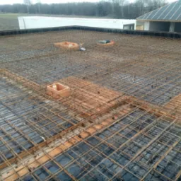 Zbrojenie stropu domu jednorodzinnego z widocznymi elementami szalunku i narzędziami, w tle budynek w stanie surowym otwartym oraz kontener.